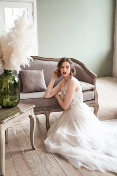 Gorgeous woman with red lips in white dress looking away while sitting on floor beside sofa
