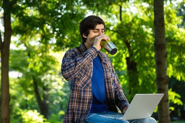 Young brunet freelancer with beard and mustache sitting with a laptop and coffee in a park.