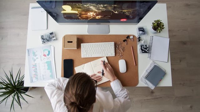 Flat lay of Caucasian businesswoman wearing white shirt and eyeglasses sitting at desk, looking at computer screen, having distance call and making notes in notebook lying on desk
