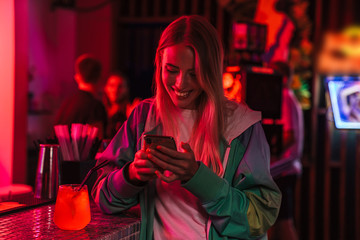 Image of young woman holding smartphone and drinking cocktail in bar