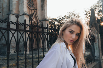 Fashionable beautiful blonde girl with loose hair, wearing white shirt, black forged fence on the...