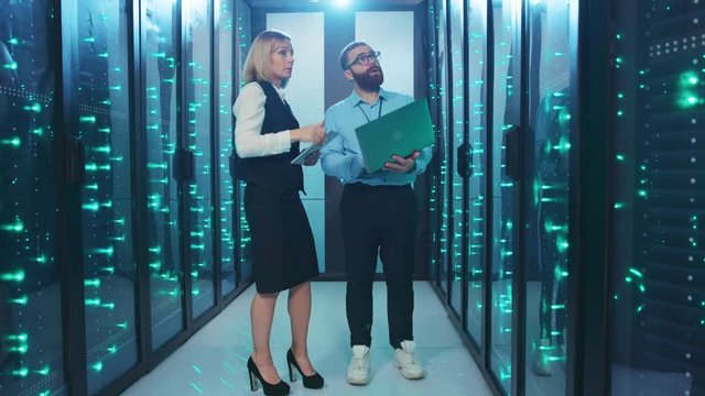 Data center IT specialists coworking in secure server room of cyberspace. Female administrator pointing at error server cabinet running diagnostics on laptop computer. Teamwork.