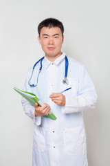 A young Asian doctor in a white coat, with a stethoscope on his neck, holds a green folder in his hands and takes notes. One on a white background.