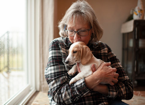 60 year old woman kissing her puppy at home sitting by window
