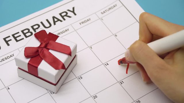 female hand with a red marker draw a heart shape on a calendar February 14 near a gift. Valentine's Day
