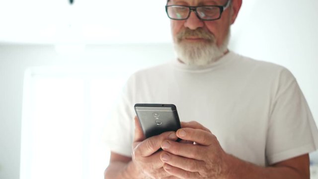 Close portrait of a gray-bearded man in glasses and a white T-shirt is using a smartphone. Smiling mature man leafing through photos in smartphone. Modern pensioner