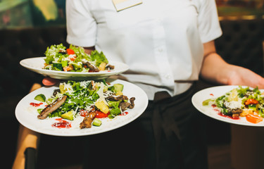 Waitress is holding fresh salad plates in her hand. Woman sets the table at the restaurant. Cafe service for birthday or wedding celebration. Different dishes on the served table.