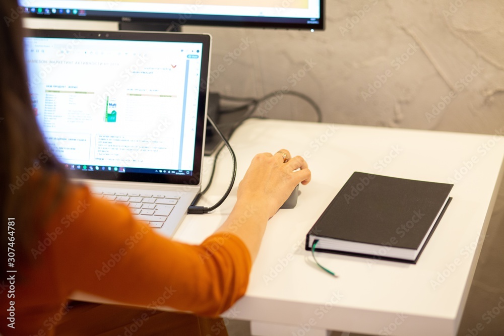 Wall mural Woman wearing an orange shirt working with a computer sitting on a desk with a black notebook on it