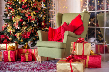Interior decoration for winter holidays: living room with white walls, sand coloured armchair and advent fir tree decorated with ribbons, knots, red and golden balls, and gift boxes. Christmas evening