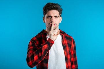 European handsome man holding finger on his lips over blue background. Gesture of shhh, secret, silence