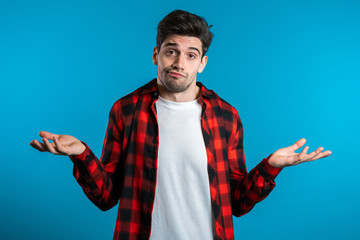 Young handsome unsure guy in red plaid shirt shrugs his arms, makes gesture of I don't know, can't...