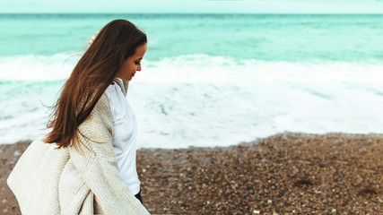 Fototapeta na wymiar A beautiful young girl walks along the seashore, a storm, hair fly apart, a gray cardigan, a sports figure, in sneakers, enjoys, autumn or winter