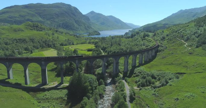 Wide aerial of Hogwart's Express (Harry Potter) steam train travelling over the Glen Finnan Viaduct in the Scottish Highlands with Loch Shiel in the background on a sunny day