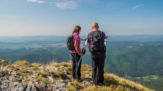 A couple is looking at a map and are standing on the top of a mountain. The weather is really fantastic.