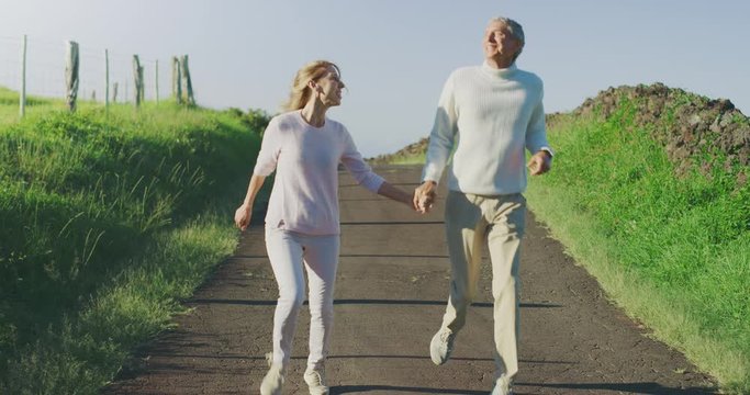 Happy retired couple having fun frolicking together on a country road, older couple skipping and running happily outdoors together on a sunny day with green fields, enjoying their retirement years