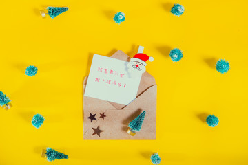 Top view festive composition with handmade stamp greeting card, santa clause pin in craft envelope with stars and many toy christmas trees on bright yellow background. Flat lay, top view. Copy space.