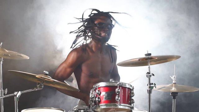 Young african man with dreadlocks sit beating on drums with sticks, with naked skin isolated over smoky background