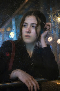 portrait of attractive brunette girl at the bus stop waiting for the bus through the glass during the rain