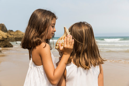 Two young girls listening to a conch shell on a beach