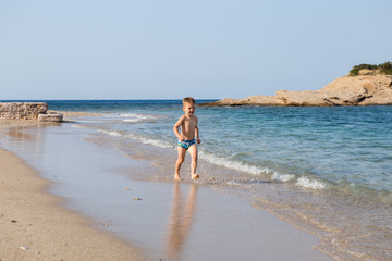 Fototapeta na wymiar A little cute boy runs along the beach and plays. Happy kid at sea. Vacation at sea with children. Greece