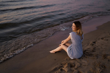 Fototapeta na wymiar Sitting: Portrait of a Beautiful blonde woman in a light blue dress on the Baltic Sea beach during sunset with vivid colors