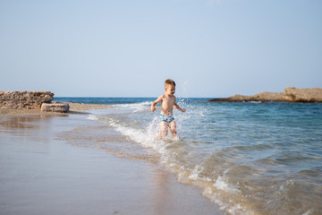 Fototapeta na wymiar A little cute boy runs along the beach and plays. Happy kid at sea. Vacation at sea with children. Greece