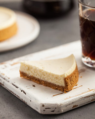 close-up of a triangular slice of cheesecake cake on a white wooden tray. In the background, a coffee pot, a mug of coffee and cheesecake.