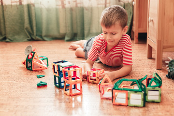 Cute boy plays with magnetic constructor in a children room laying on a cork floor