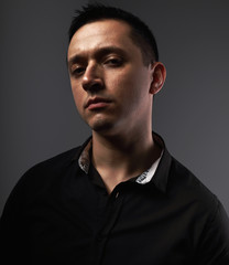 Sexy young man looking serious in black shirt on grey studio dark shadow background. Closeup portrait.