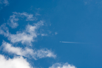 A distant airplane leaving an horizontal steam tail in blue sky with some clouds to its left