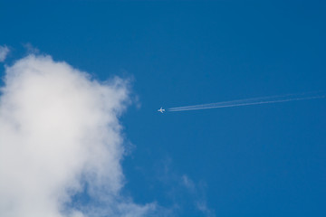 A distant airplane leaving a steam tail in a cloudy and blue sky crossing from right to left