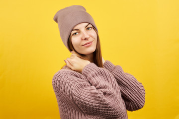 Fototapeta na wymiar happy smiling girl in sweater and warm hat isolated on an yellow background. one person. outer space. hands on the neck. winter season.