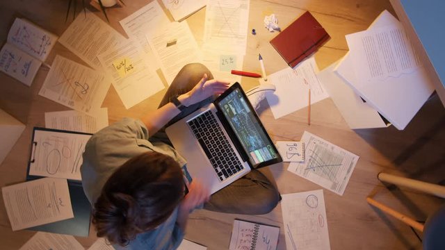 Flat lay of young woman sitting on floor surrounded with documents and charts with laptop on her knees and working on it