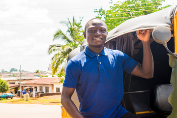 cheerful african man standing next to his tuk tuk taxi smiling