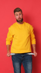 Fototapeta premium Young man is showing his pocket out. Man in yellow blouse on red background. Empty pockets. 