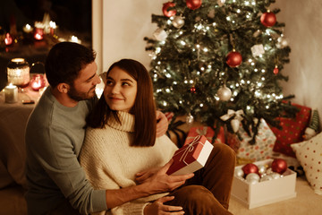happy couple sitting near xmas decoration on background; man giving gift box to woman and they look at each other