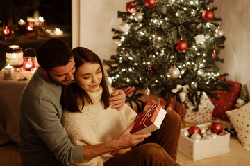 happy couple sitting near xmas decoration on background; man giving gift box to woman