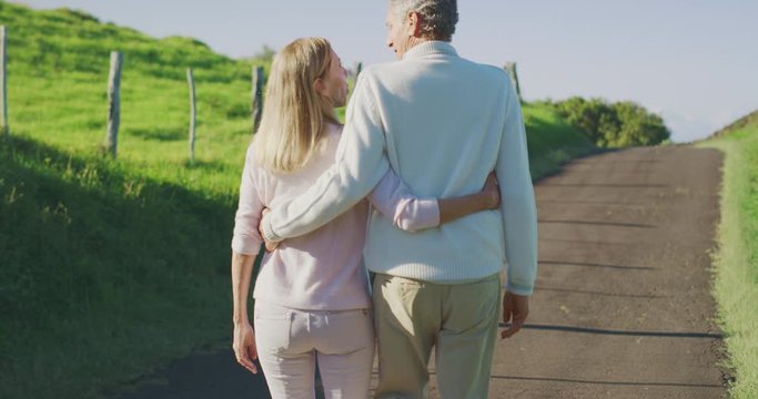 Happy elderly couple taking a walk outdoors together on a sunny country road with green pasture fields, older mature couple enjoying the golden years of retirement