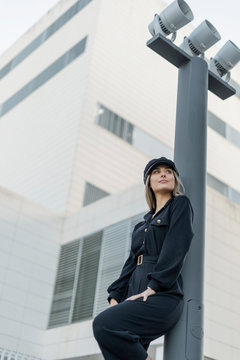Young blond businesswoman wearing black sailor's cap and looking sideways, leaning on a lamp pole, low angle view