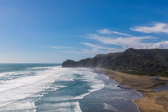 Scenic view of cliff by sea against blue sky at Piha Beach, Auckland, New Zealand