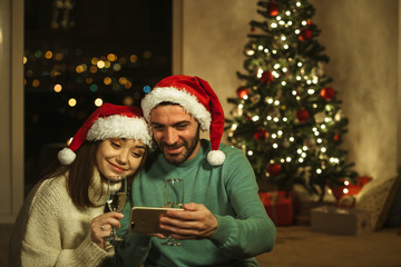 Obraz na płótnie Canvas happy couple drinking champagne with santa hat and watching video on smart phone; christmas celebration concept