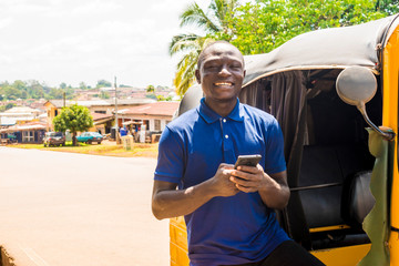 cheerful african man standing next to his tuk tuk taxi smiling and using his smart phone