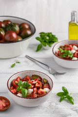 Two bowls with caprese salad, fresh black prince tomatoes in enamel pan, olive oil bottle, bowl with basil and a fork on white wooden table. Organic and vegetarian food concept