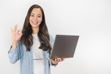 Portrait of young asian girl holding laptop computer and showing ok gesture isolated over white background
