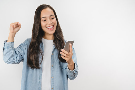 Happy Satisfied Asian Girl In Casual Wear Holding Mobile Phone Isolated Over White Background