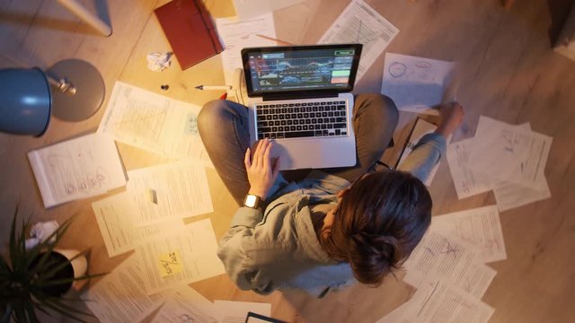 Pan shot of young woman sitting on floor surrounded with different papers and documents with laptop on her knees and working in the late evening