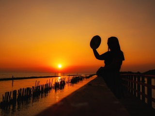 Fototapeta premium A woman silhouette standing Selfie on wooden pier at the sea with beautiful cloudy sunset.