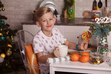 Small boy having white cup of tea or milk holding and drinking at home by the table. Beautiful boy with a big candy sits at the kitchen table. Christmas holidays, new year.