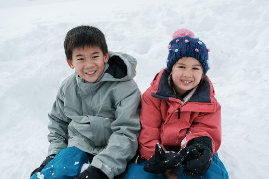 Happy Mixed Race Asian Boy And Girl, Siblings Smiling And Sitting On White Snow In Japan, Family Holidays, Preteen Travel