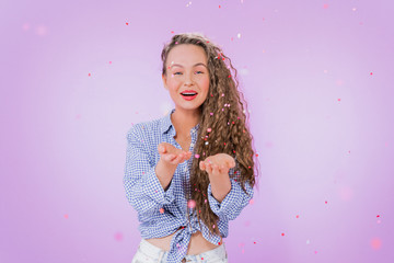 A beautiful curly-haired girl catches colored paper confetti on her palms. Portrait of a girl in a rain of confetti on an isolated purple background.